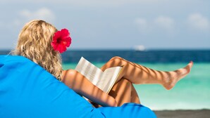 Iemand leunt ontspannen op een blauw kussen aan het strand, leest een boek, met een rode bloem in het haar, en de oceaan op de achtergrond.