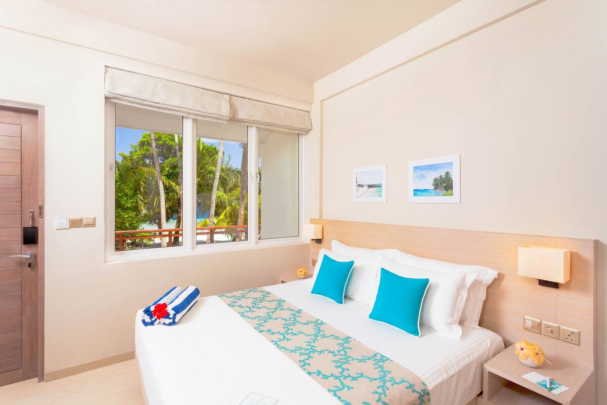 A hotel room with a bed, a window with a view of palm trees, and a small bedside table with a lamp.