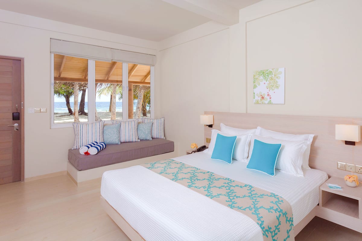 A bedroom with a large bed, a window with a view of palm trees and the sea, a bedside table with a lamp, and a small nightstand.