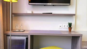 Una habitación de hotel moderna con un televisor de pantalla plana montado en la pared, un escritorio pequeño con una planta en maceta y un taburete verde brillante.