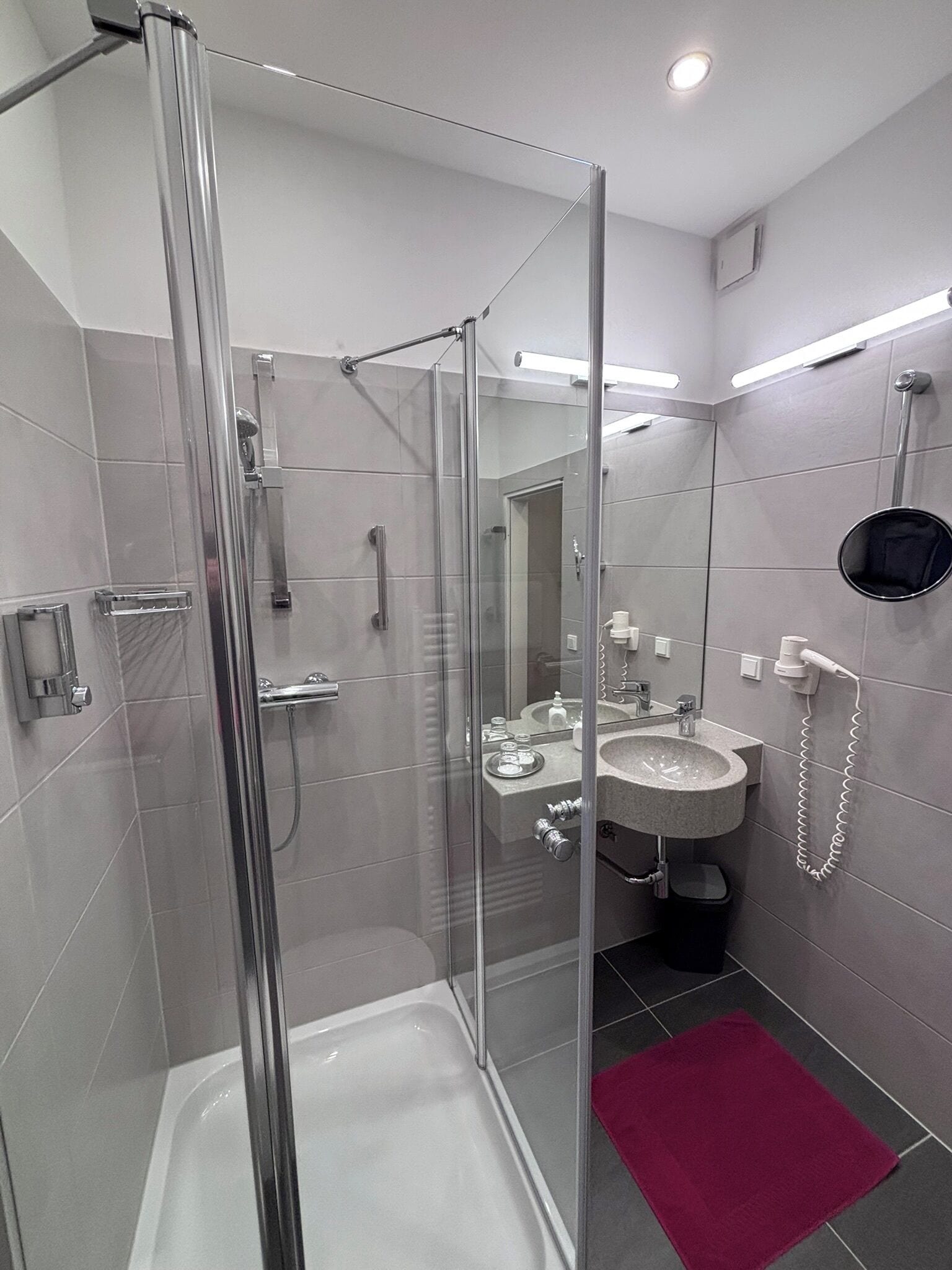 A modern bathroom with a glass-enclosed shower, a white bathtub, a sink with a mirror, and a red bath mat.