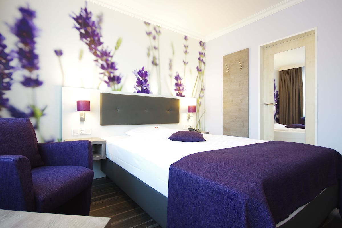 A hotel room with a large bed, two armchairs, a nightstand, a wall-mounted lamp, and a mirror.