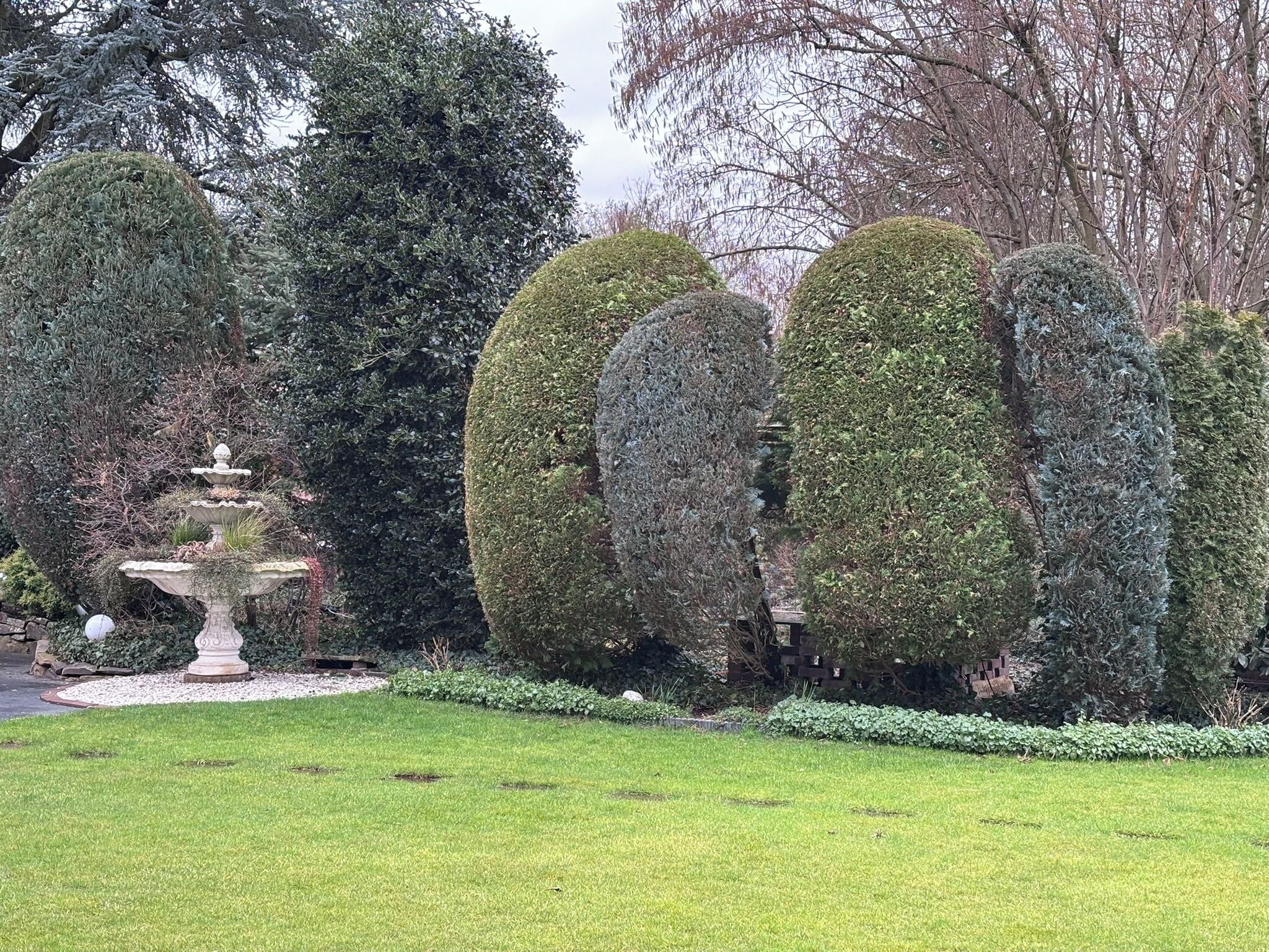 A well-maintained garden with a tiered stone fountain, neatly trimmed hedges, and a variety of trees.