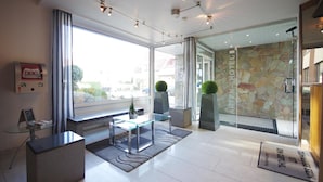 Un hall d’hôtel moderne avec un comptoir d’accueil, une table basse en verre et un mur en pierre portant le nom de l’hôtel.