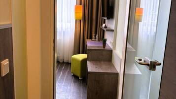 A hotel room with a wooden floor, a green ottoman, a small table with a lamp, and a TV on the wall.