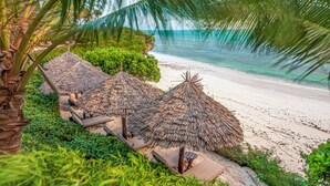 A beach with thatched-roof structures, palm trees, and a clear view of the ocean. - Zawadi Hotel Zanzibar - All Inclusive (Dongwe)