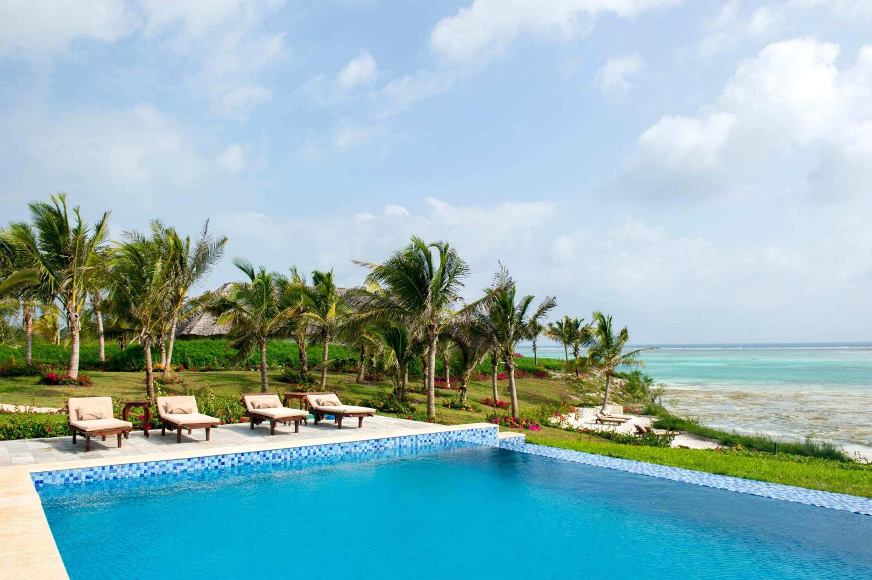 Zona junto a la piscina con sillones de descanso y palmeras, con vista a una playa y al océano.