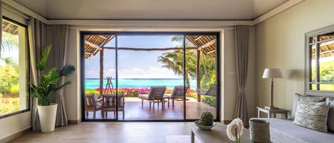 A room with a large sliding glass door leading to a balcony with a view of the ocean, a potted plant, a sofa, a lamp, and a table with decorative items.