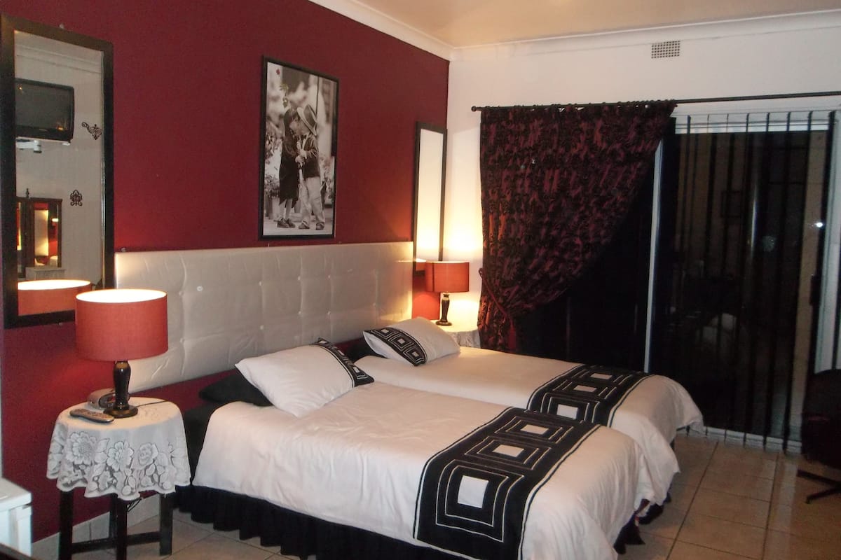 Ein Hotelzimmer mit zwei Betten, einer roten Wand, einem gerahmten Bild, einem Spiegel und einem Vorhangfenster.