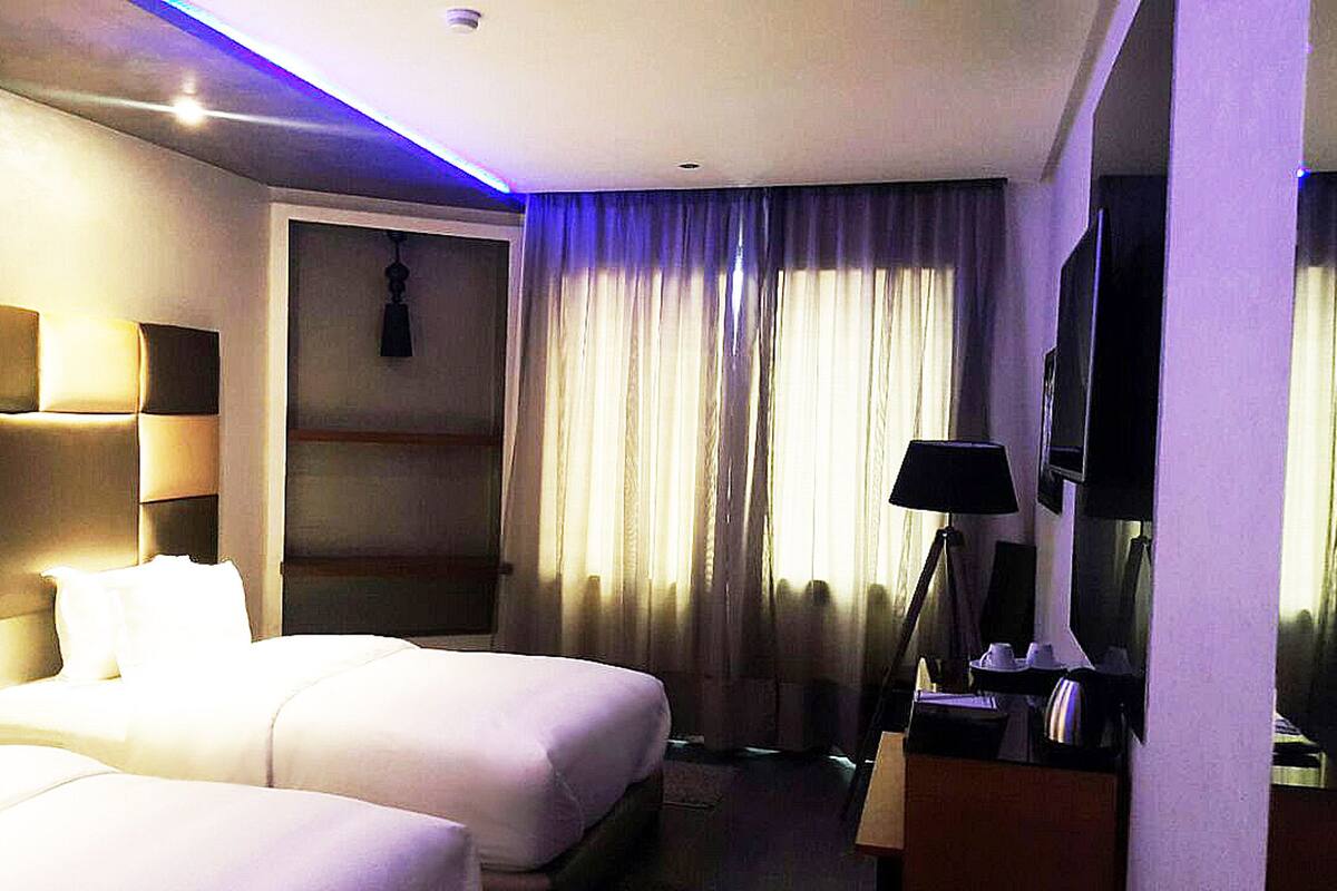 A hotel room with two beds, a desk with a lamp, a TV, and a large window with curtains.