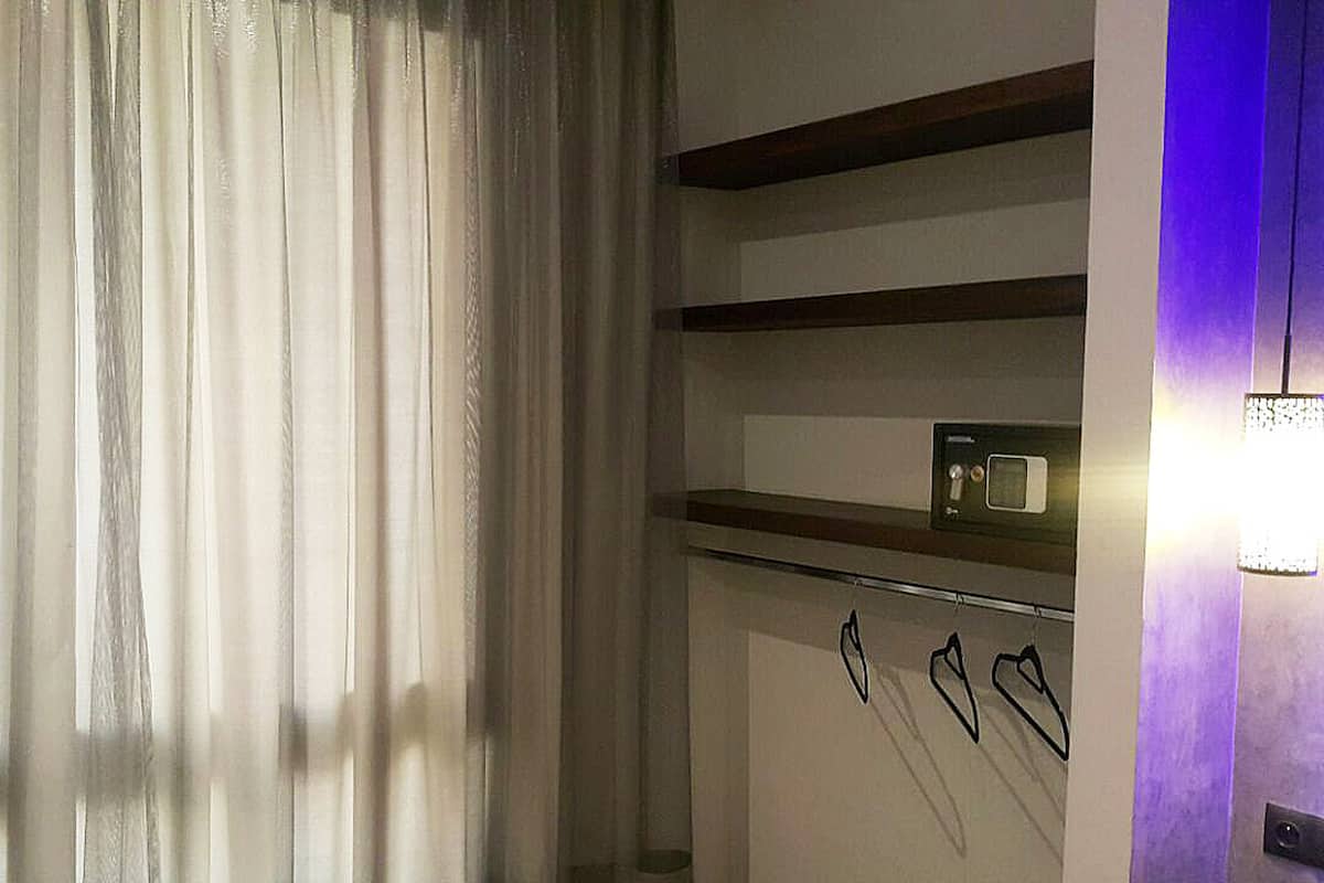 A room with a window covered by sheer curtains, a wall-mounted shelf with a small item, and a wall-mounted light fixture.