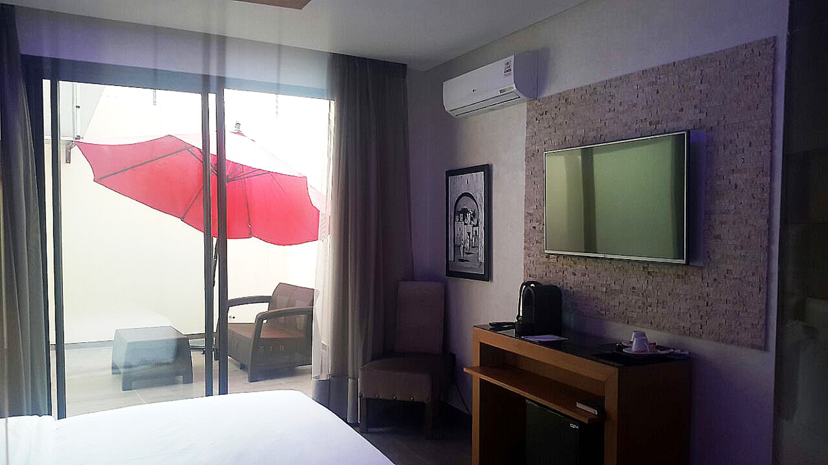 Une chambre d’hôtel avec un grand lit, un téléviseur fixé au mur, un bureau avec une chaise et une fenêtre avec un parapluie rouge.