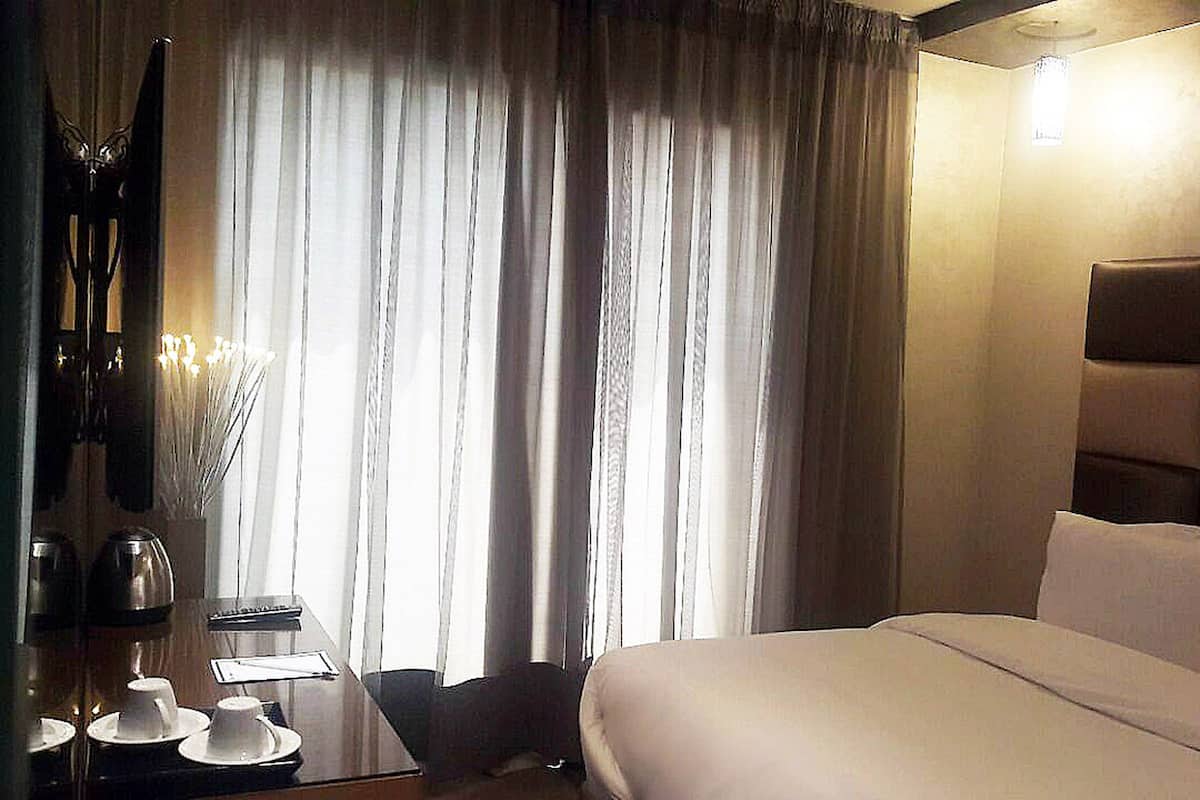 A hotel room with a large bed, a desk with a lamp, a chair, and a window with curtains.