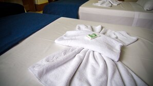 Habitación de hotel con dos camas, una con cubrecama azul y la otra con cubrecama blanca. Una toalla blanca está colocada en la cama con una etiqueta verde.