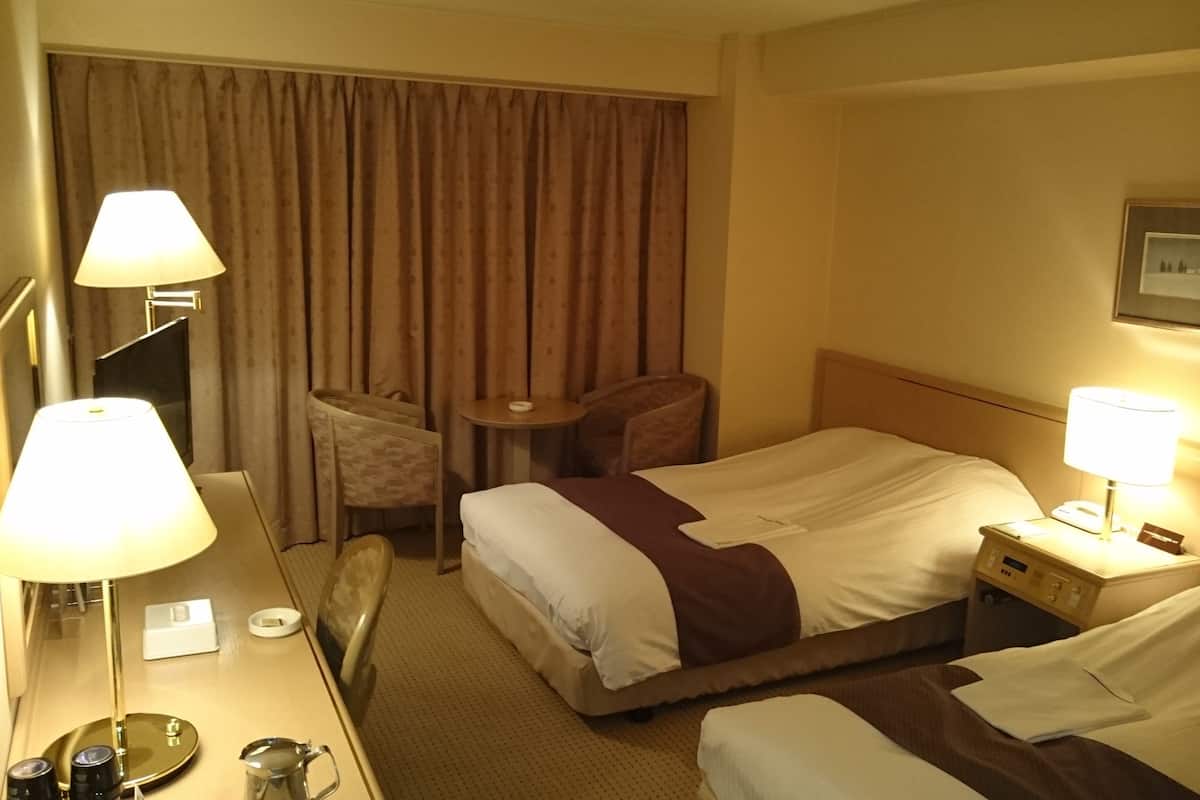 Habitación de hotel con dos camas, un escritorio con lámpara, una silla, una mesita y una cortina.