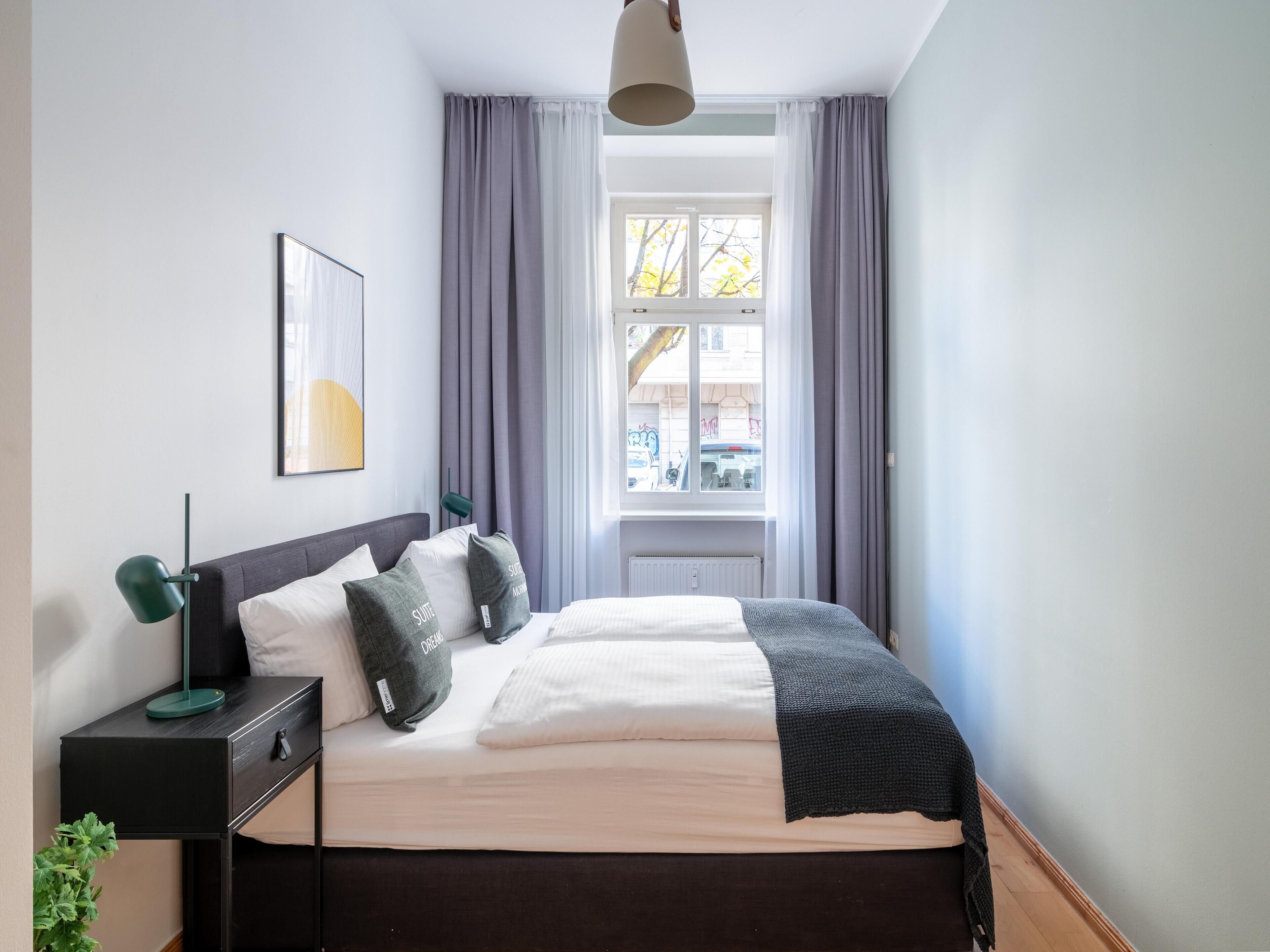 A neatly arranged bedroom with a bed, bedside table, lamp, and a window with curtains.