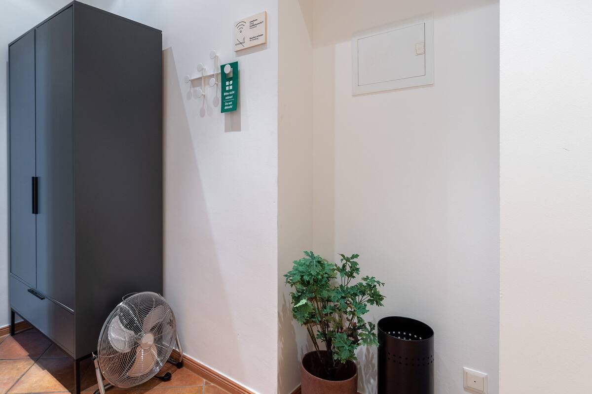 A room with a grey wardrobe, a standing fan, a potted plant, and a trash bin.