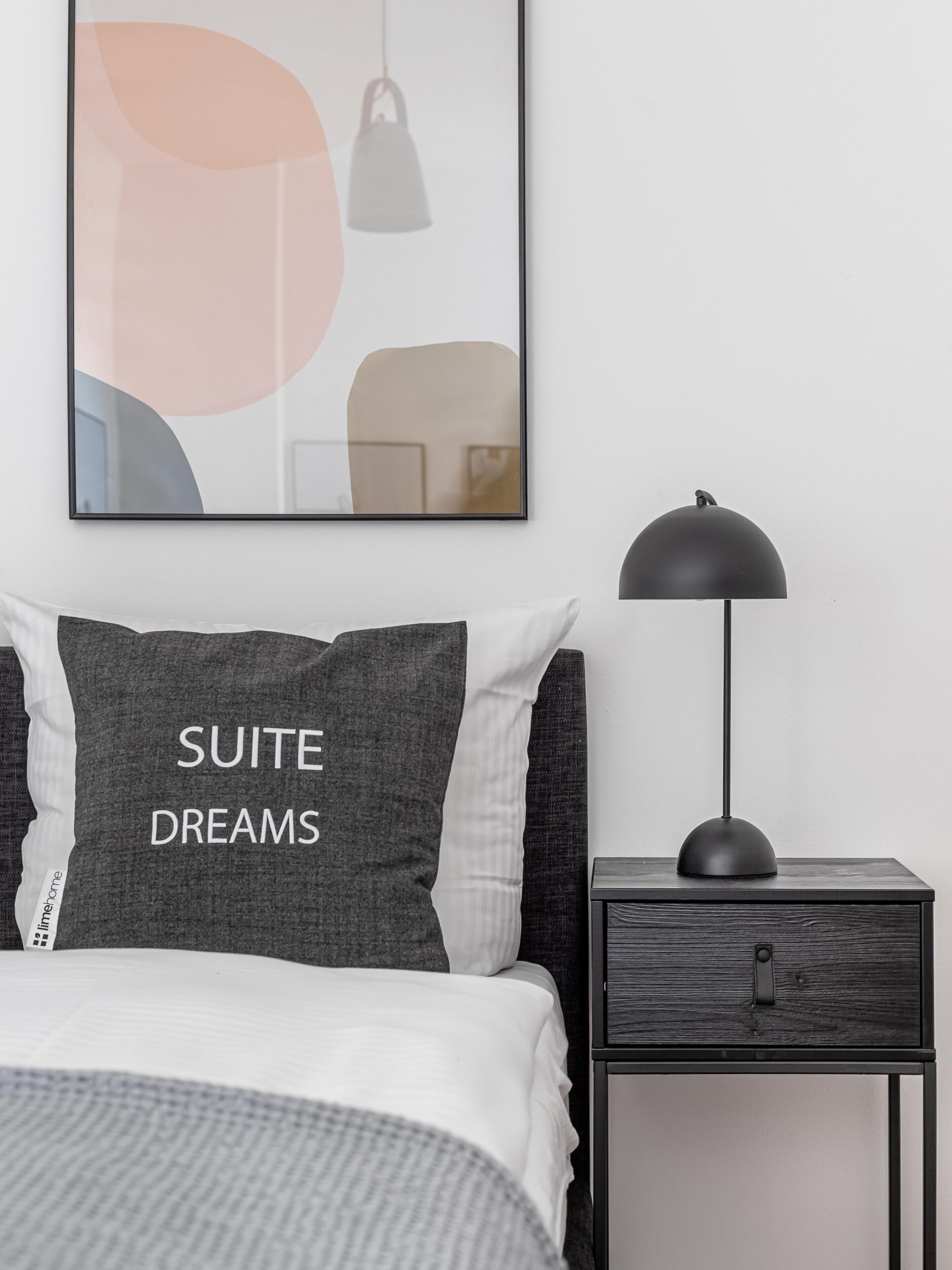 Un dormitorio ordenado con cama, una mesita de noche con lámpara y una obra de arte abstracta enmarcada en la pared.