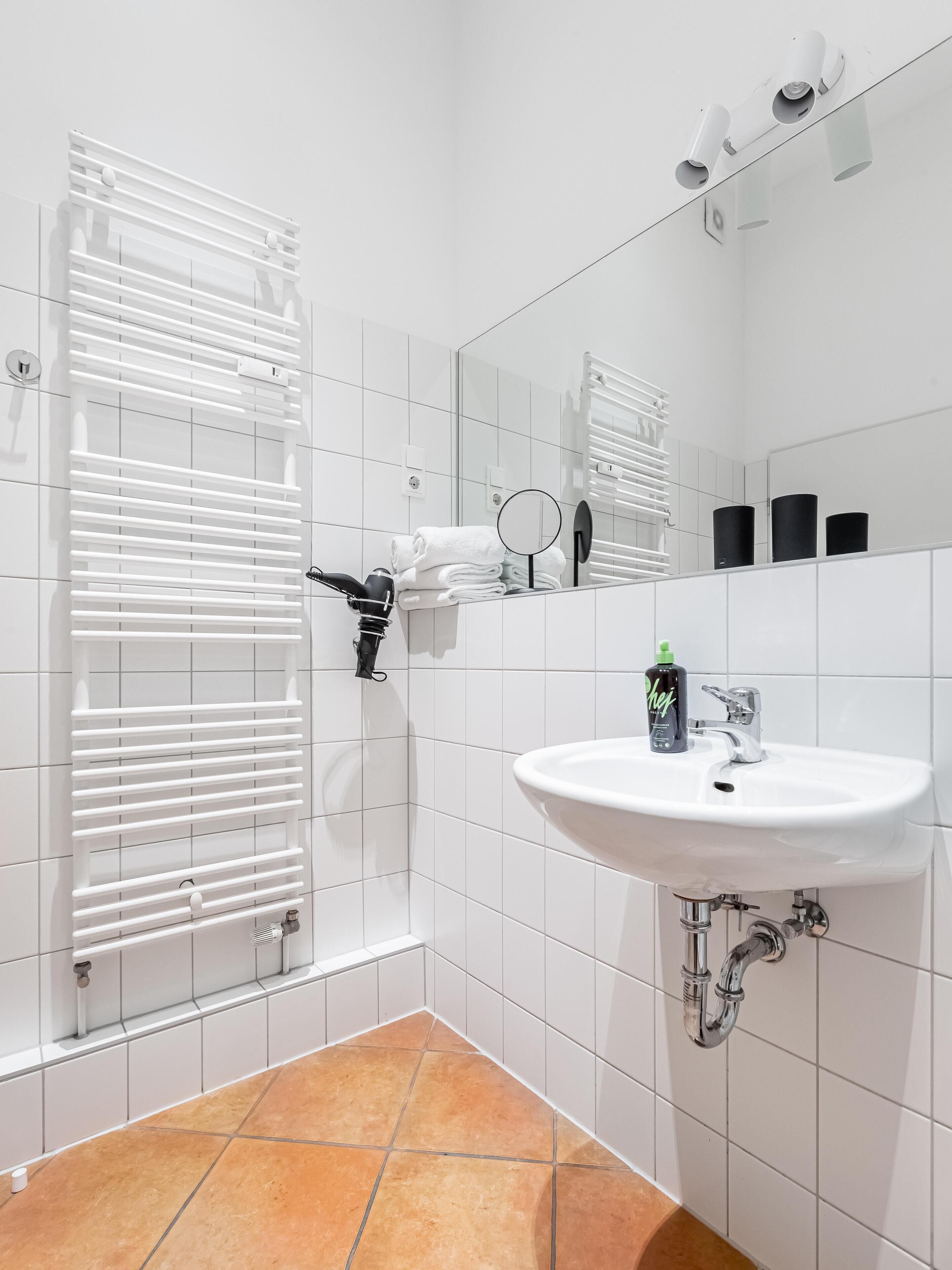 A modern bathroom with a white tiled floor, a white sink with a chrome faucet, and a towel rack with folded towels.