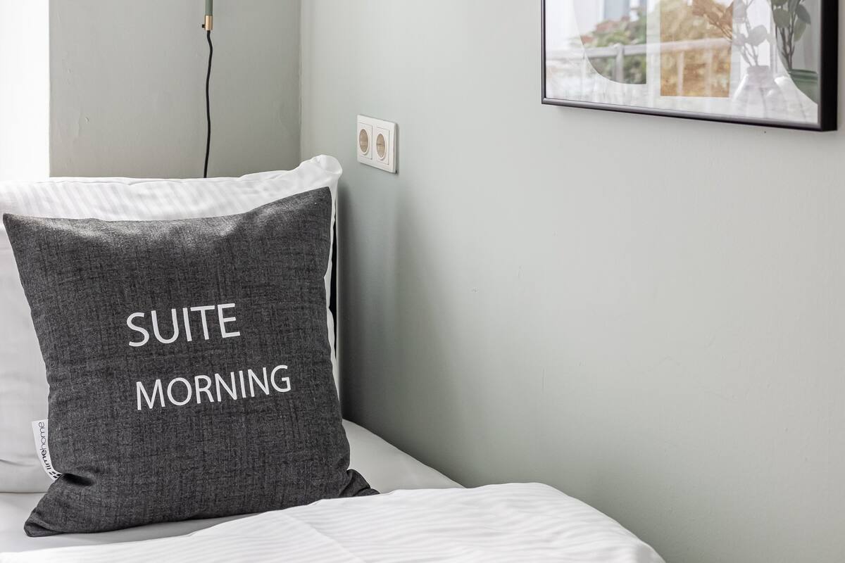 A bedroom with a bed, a grey pillow with "SUITE MORNING" text, a wall-mounted lamp, and a framed picture reflecting a building and plants.