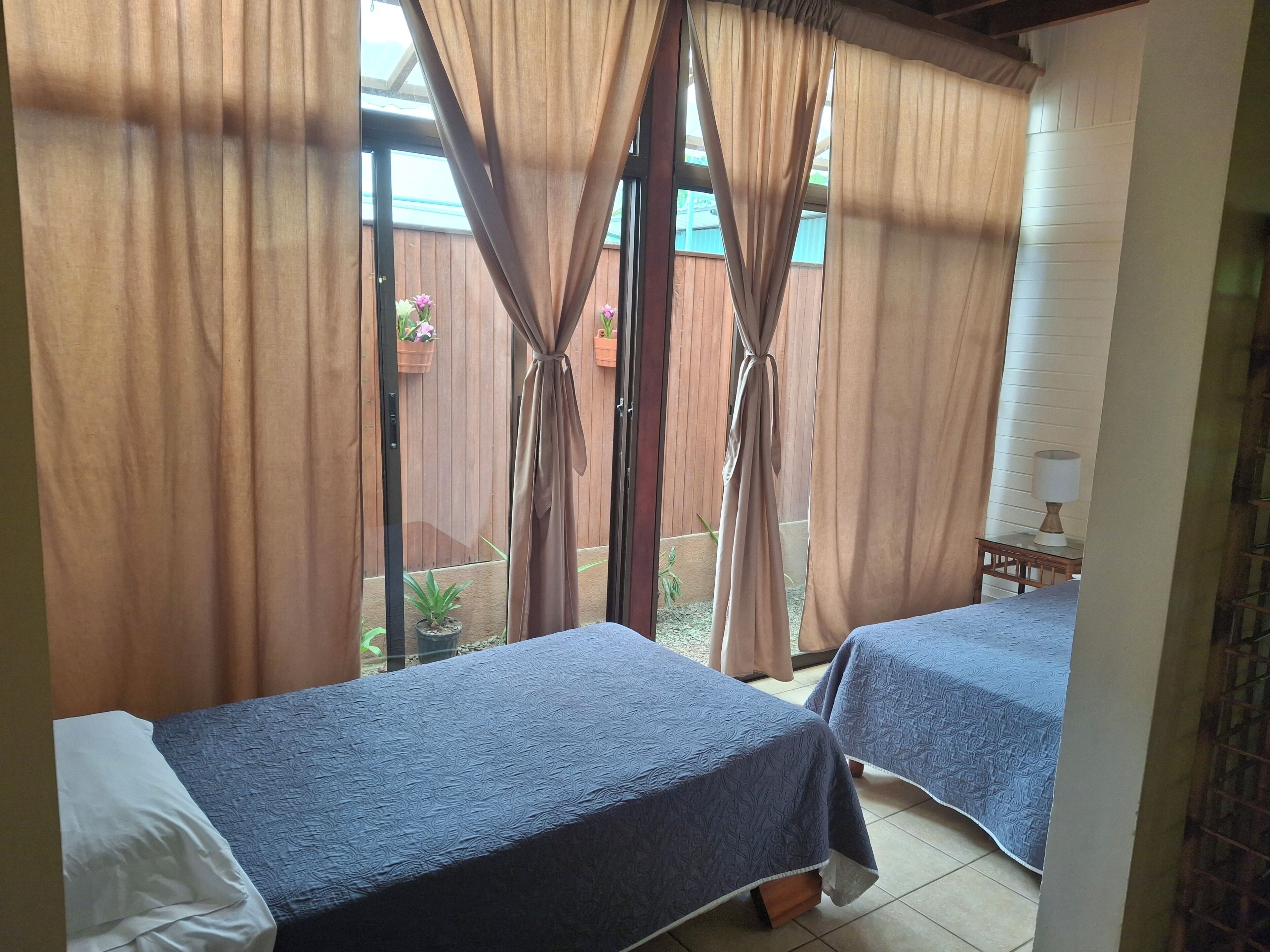 A room with two beds, a window with curtains, a small table with a lamp, and a view of a wooden fence and plants outside.