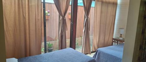 A room with two beds, a window with curtains, a small table with a lamp, and a view of a wooden fence and plants outside.
