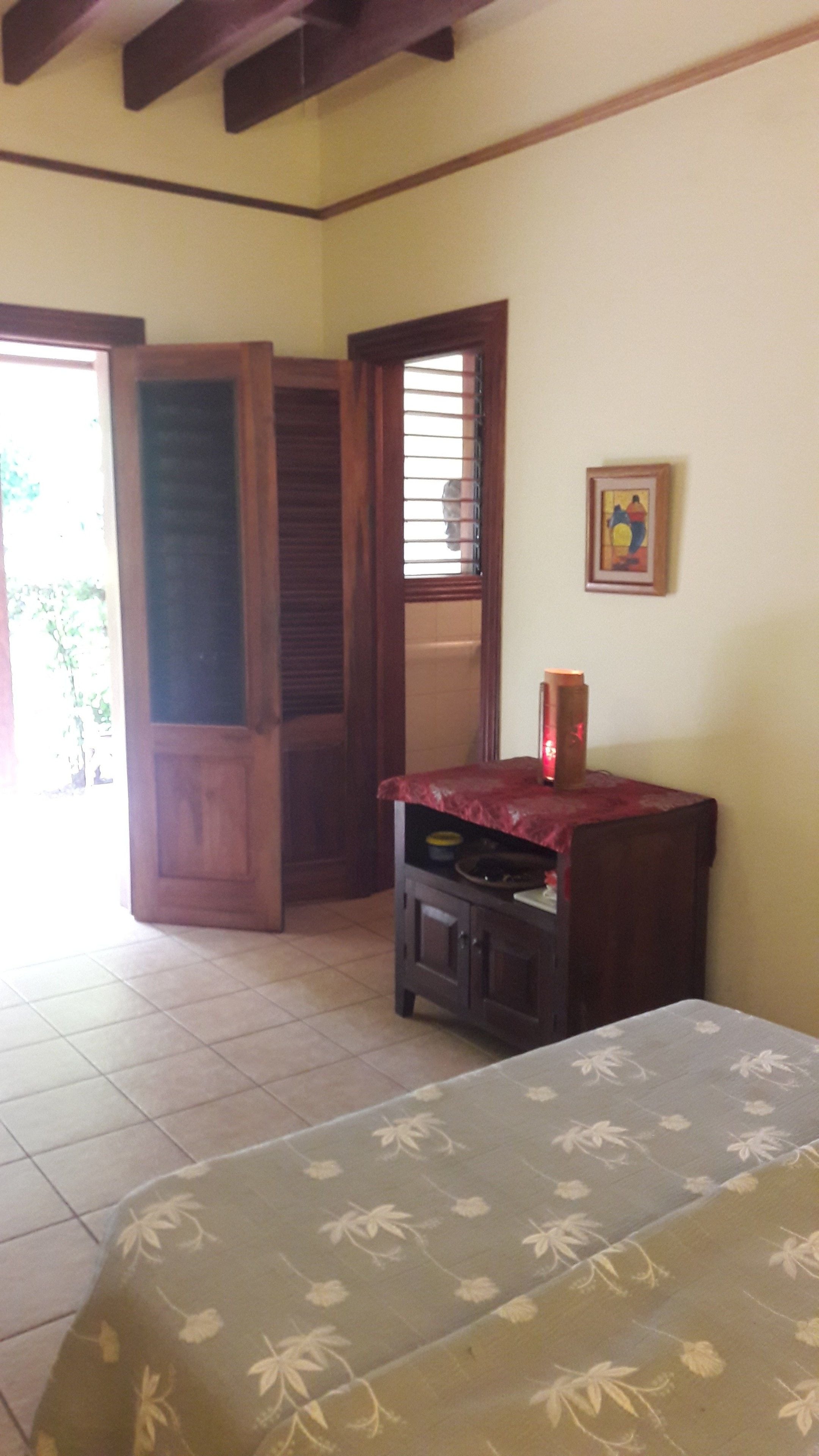 A room with a bed, a small table with a red cloth, a framed picture on the wall, and a door leading to another room.
