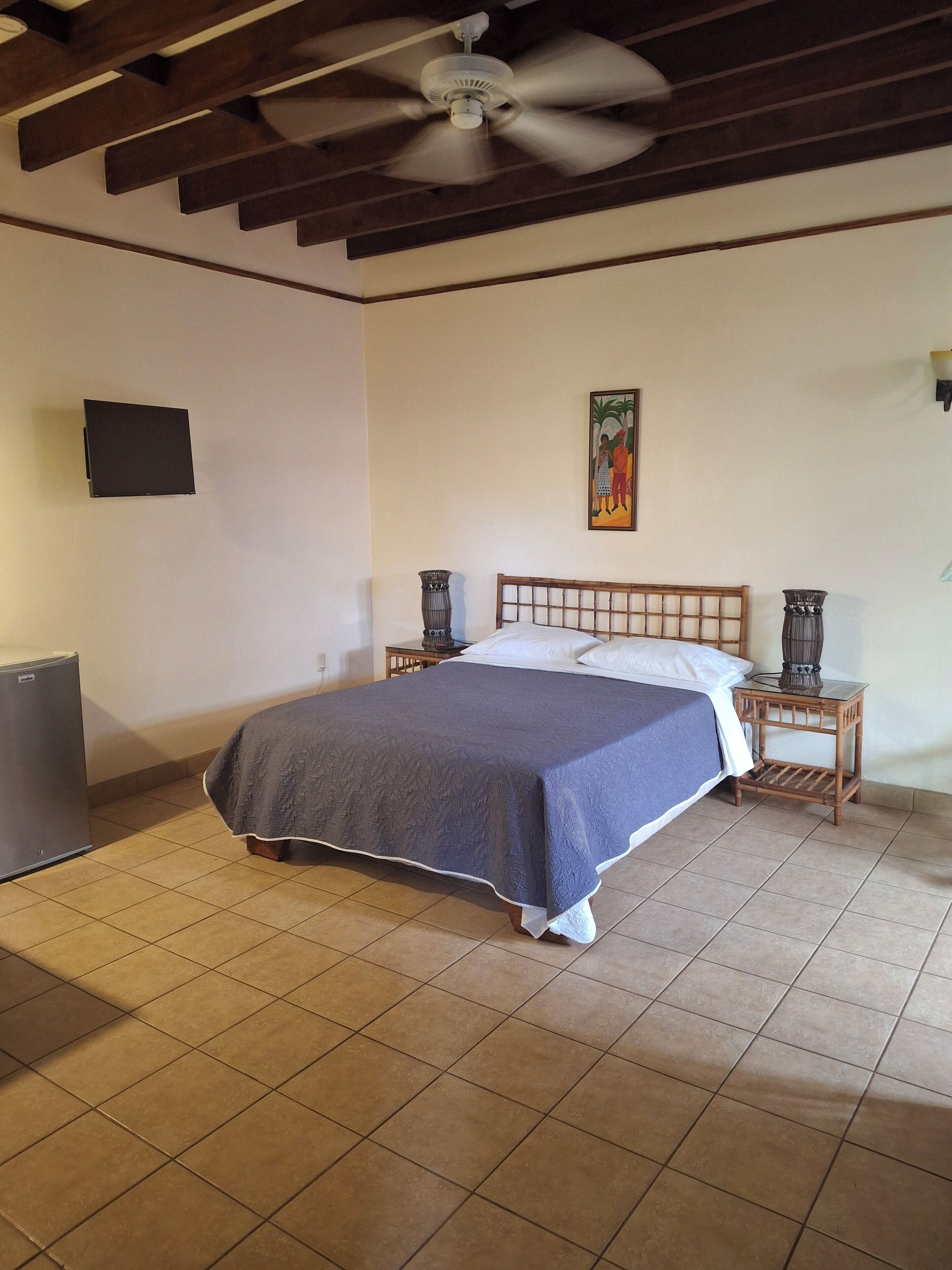 Ein Schlafzimmer mit einem Bett, einem Deckenventilator, einem Gemälde und einem kleinen Tisch mit einer Lampe.