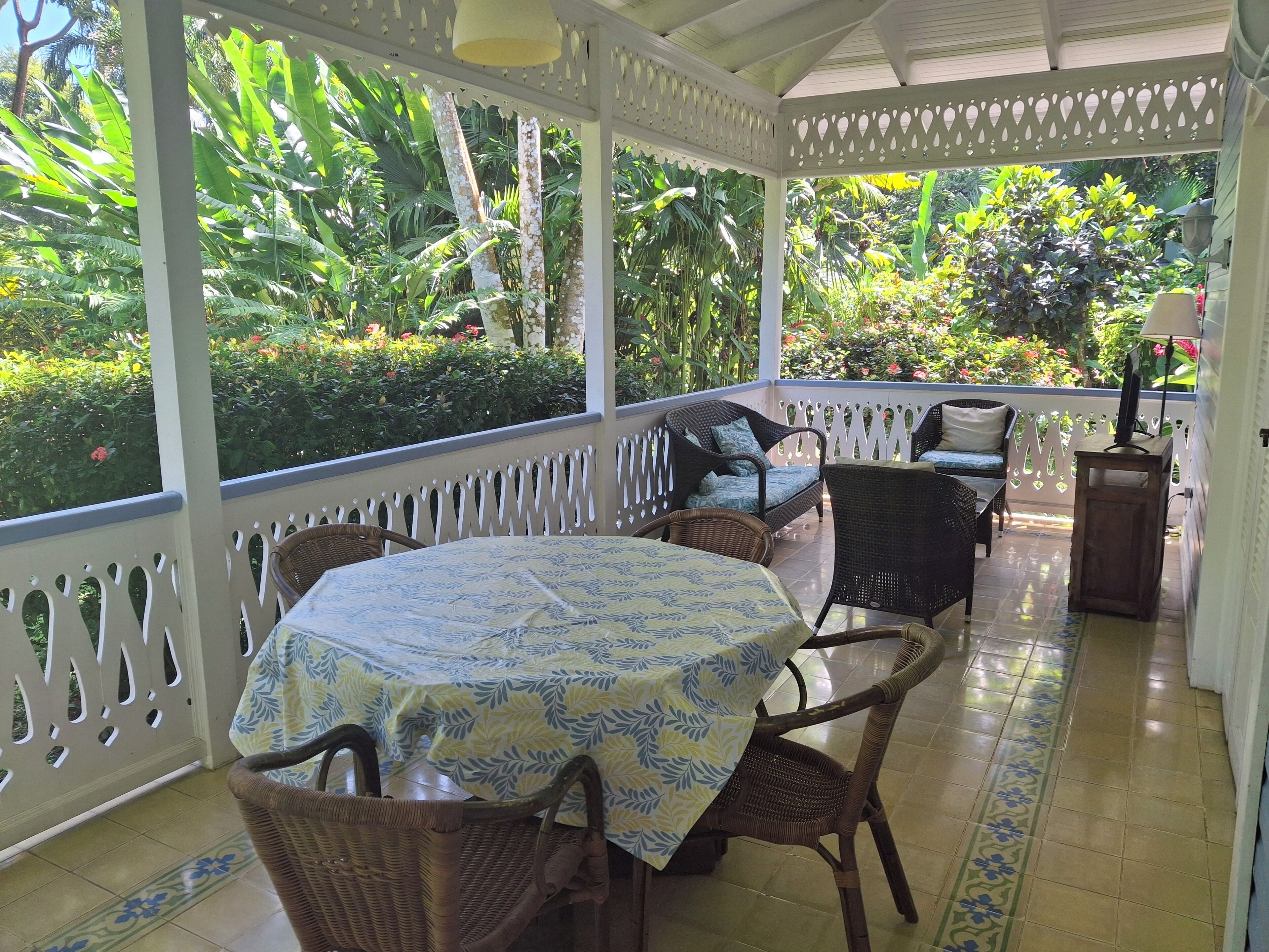 A covered outdoor seating area with wicker furniture, a table with a patterned tablecloth, and a view of lush greenery.