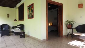 A room with a tiled floor, a bed with a white bedspread, a chair, a small table with a plant, and a wall with a framed picture.