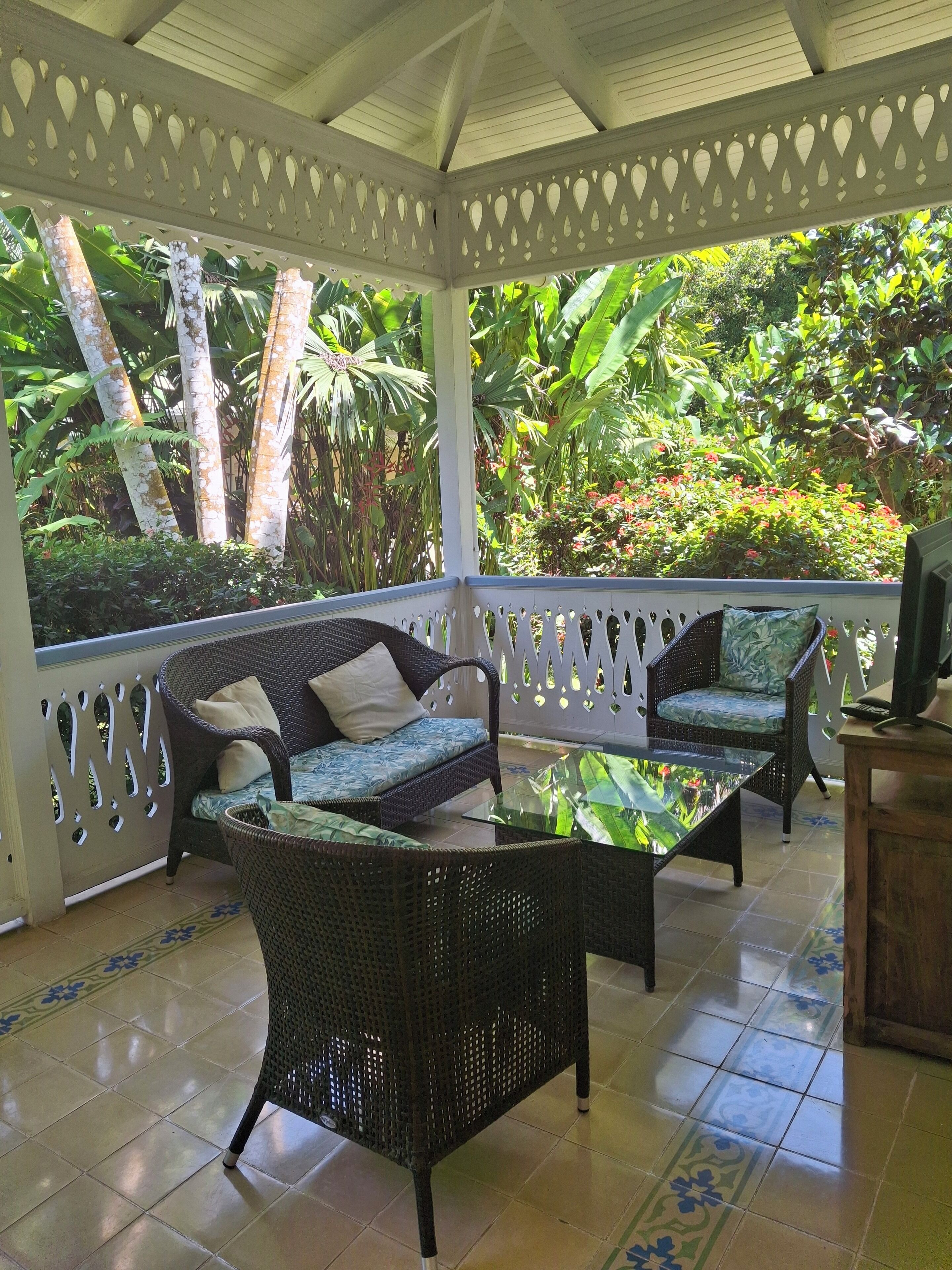 A covered outdoor seating area with wicker furniture and a glass table, surrounded by lush tropical plants.
