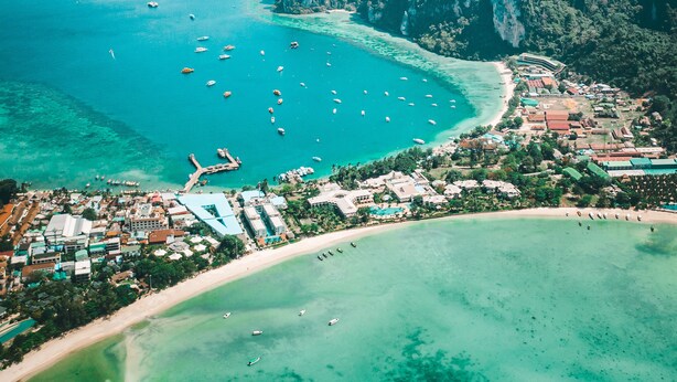 An aerial view of a coastal area with clear turquoise waters, a beach, and a cluster of buildings. - PP Princess Pool Villa (Ko Phi Phi)