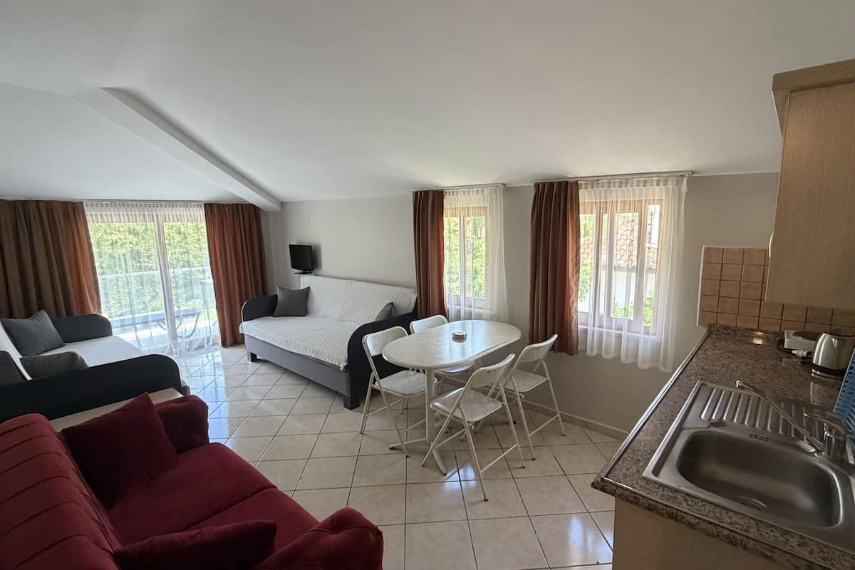 A hotel room with a bed, a sofa, a dining table with chairs, a kitchen area with a sink, and large windows with curtains.