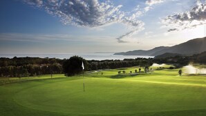Un parcours de golf avec un mât de drapeau, des arbres et une vue lointaine sur la mer et les montagnes.