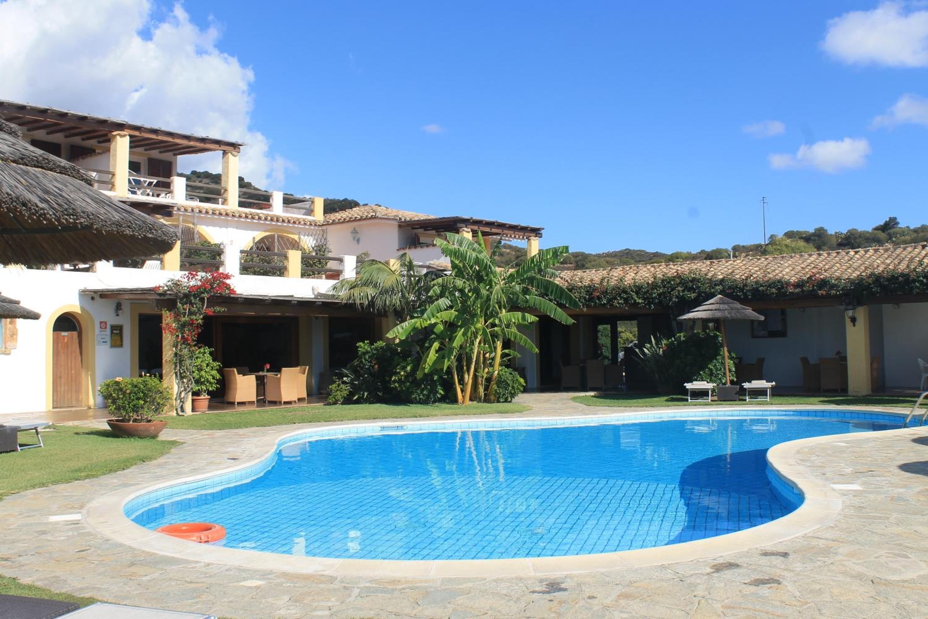 A swimming pool with a curved edge, surrounded by a stone patio. There are lounge chairs and a table with a thatched roof.