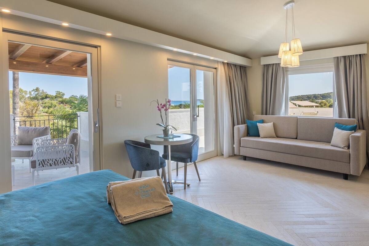 Een hotelkamer met balkon, een bank, een tafel met stoelen en een bed met een blauwe handdoek.