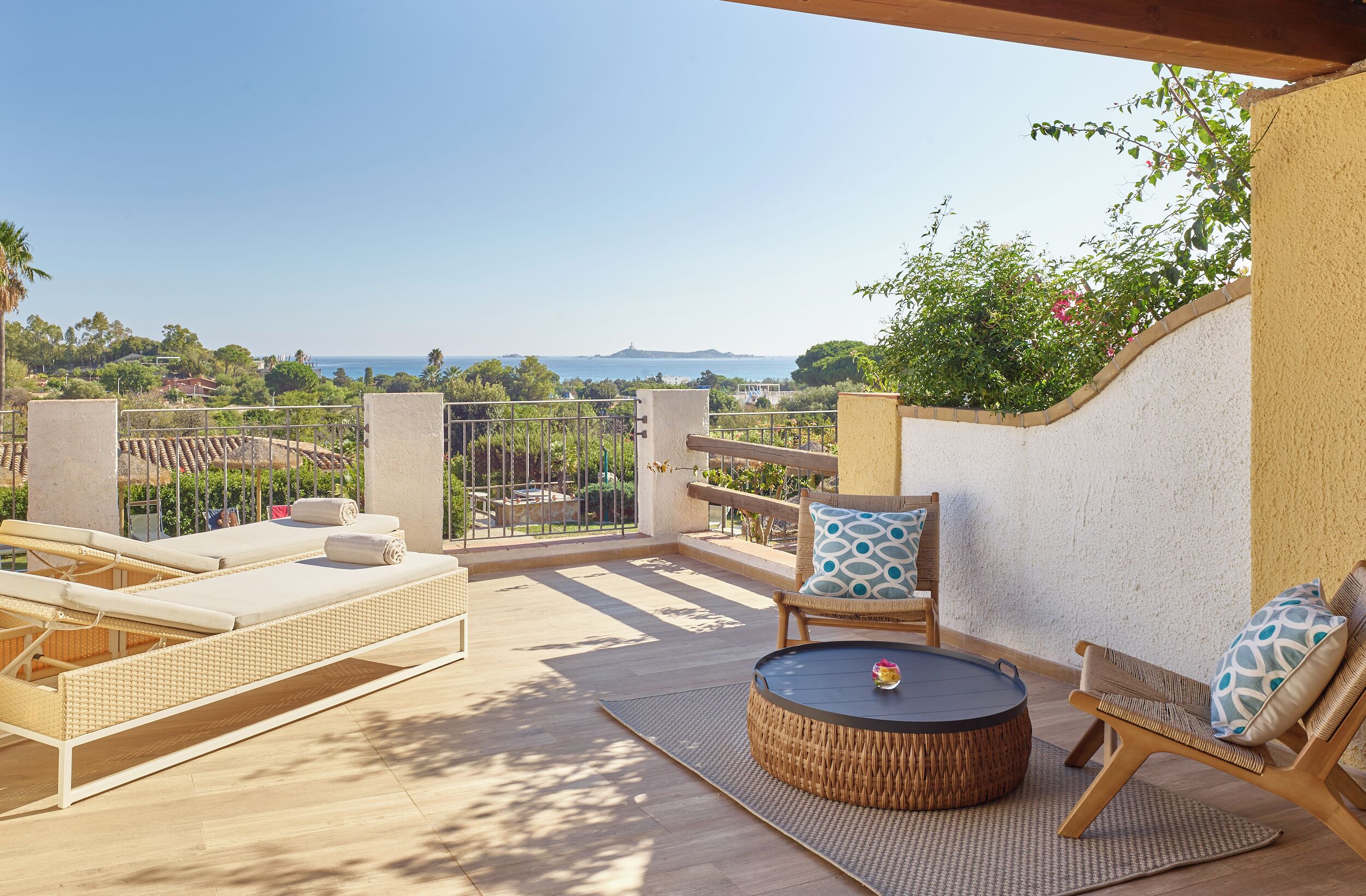 Une terrasse extérieure ensoleillée, dotée de chaises longues, d’une table ronde et offrant une vue sur la mer et la verdure.