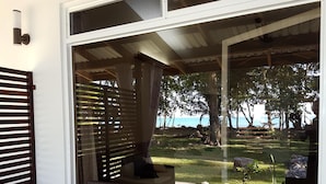 Une pièce dotée d’une grande fenêtre donnant sur une plage. On y trouve un canapé en osier et une porte en bois avec des lattes horizontales.