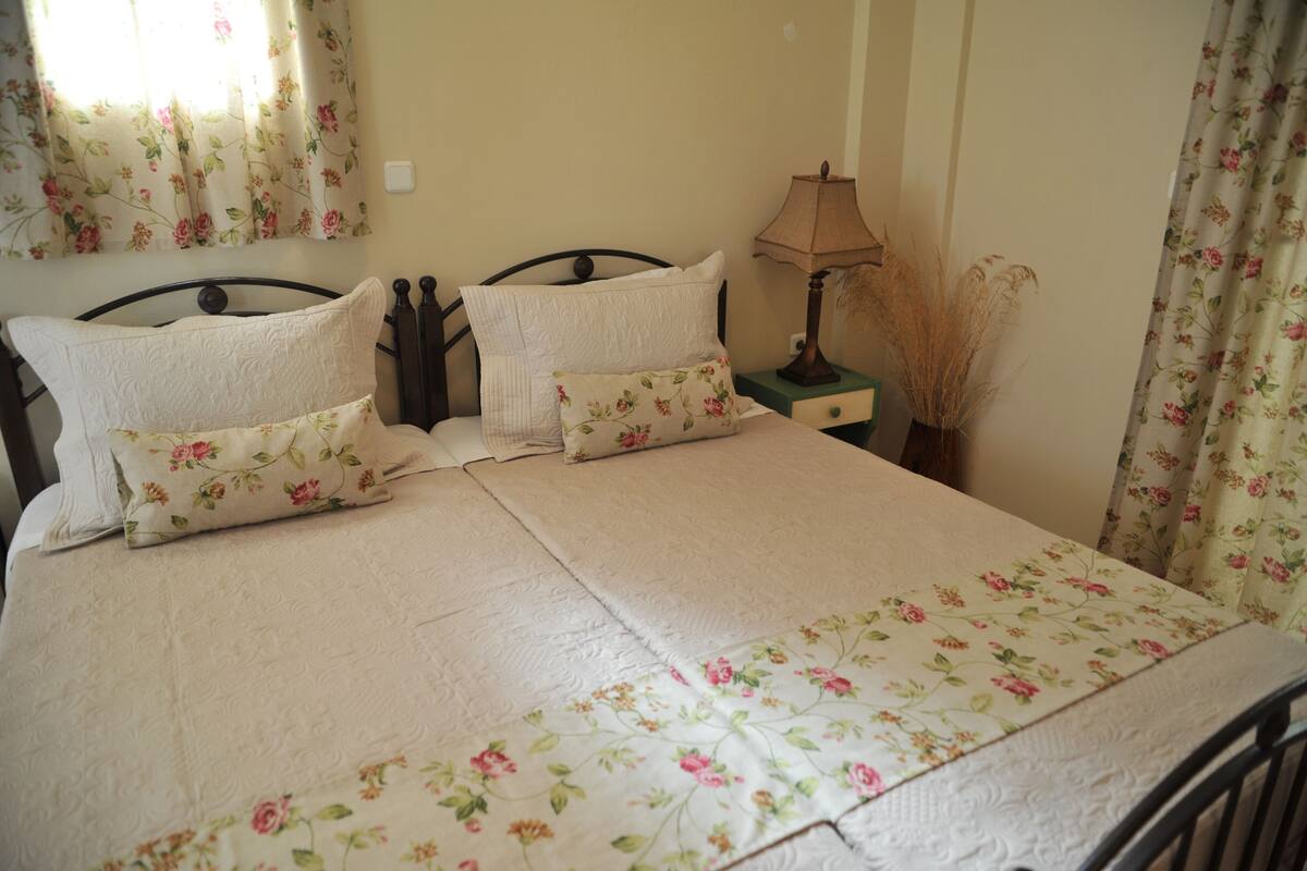 A neatly made bed with floral bedding, a bedside table with a lamp, and a window with floral curtains.