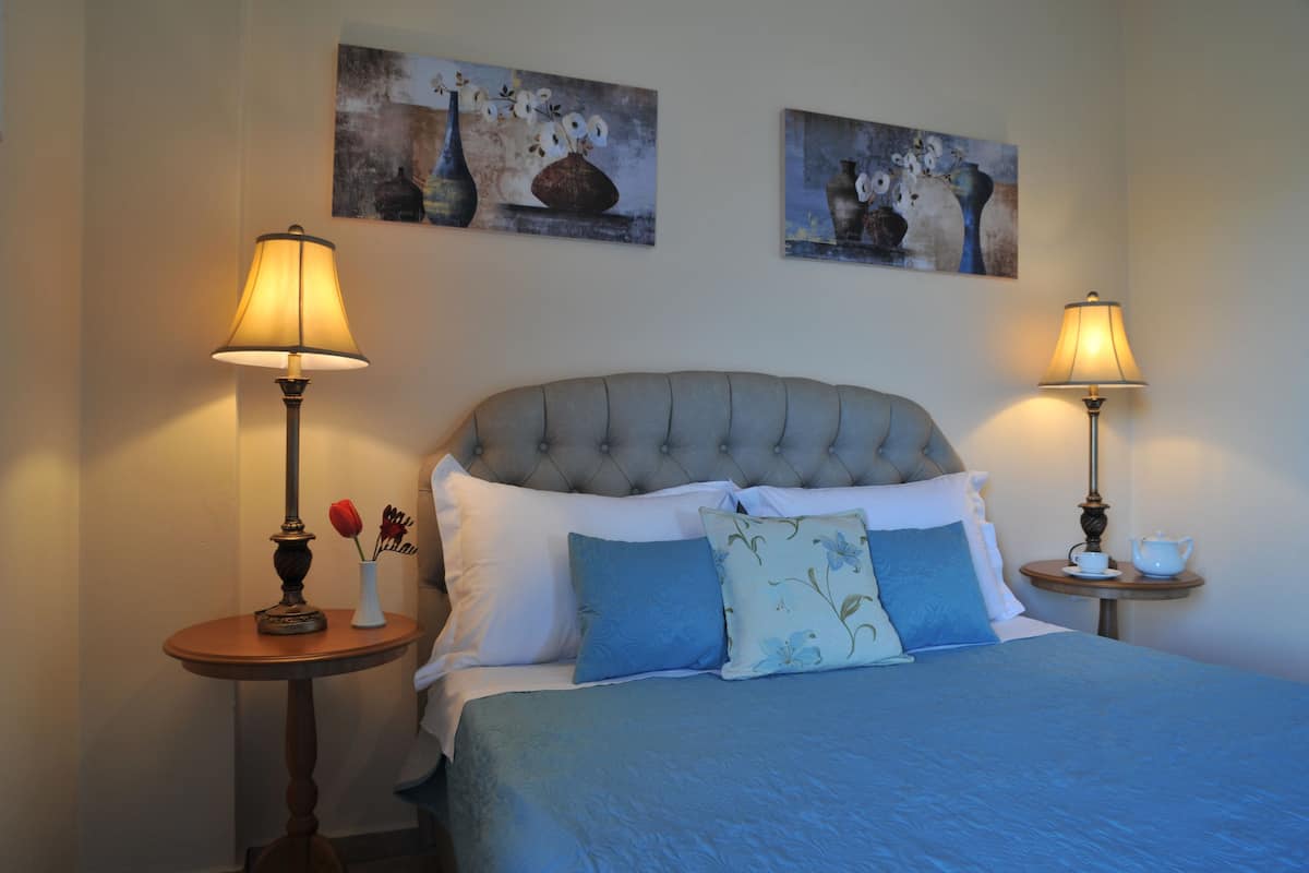 A bedroom with a tufted headboard, a bed with blue bedding, two bedside tables with lamps, and two paintings on the wall.