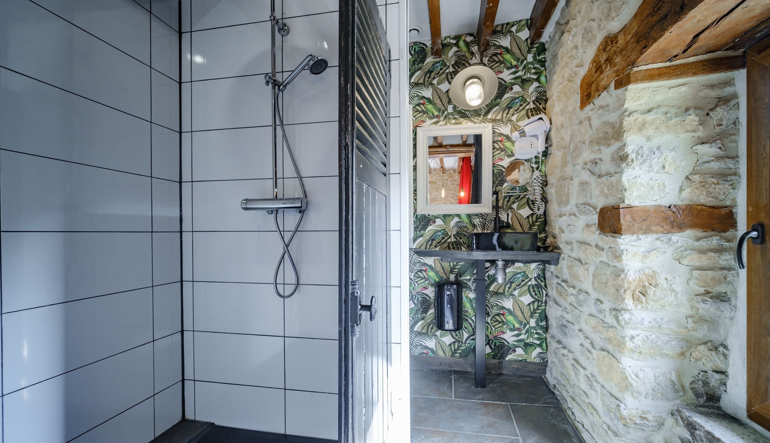 A bathroom with white tiled walls, a shower with a handheld showerhead, a sink with a mirror, and a stone wall with a wooden door.
