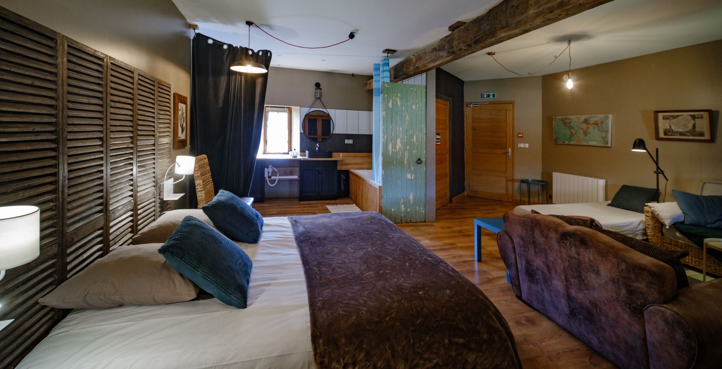 A cozy room with a bed, a sofa, and a chair, wooden beams on the ceiling, a kitchen area, and a bathroom with a towel rack.