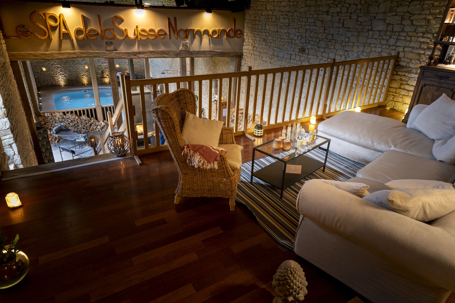 A cozy lounge area with a wicker chair, a white sofa, and a glass coffee table. A stone wall and a sign reading "Le SPA de la Suisse Normande" are visible in the background.