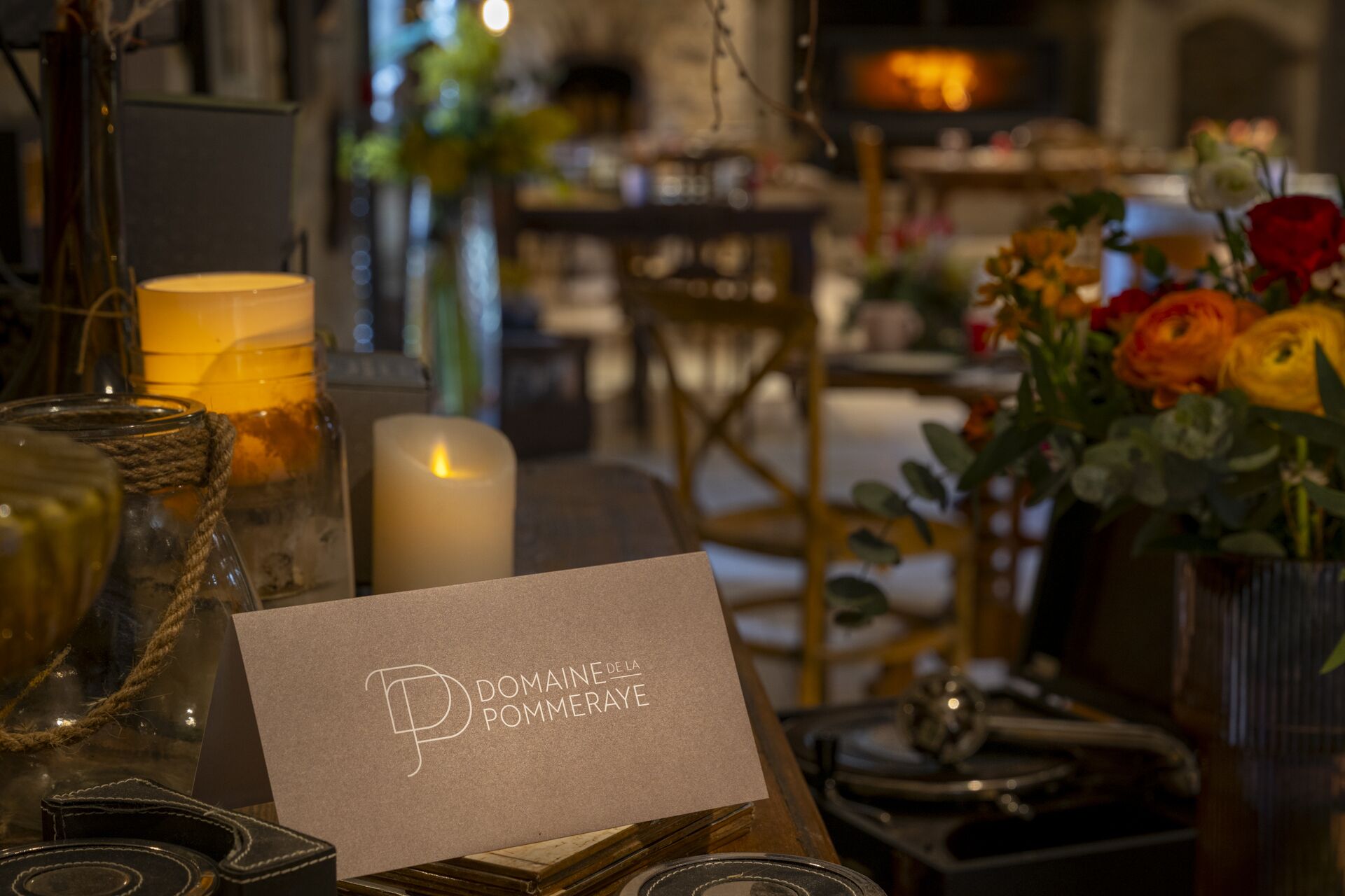 Una mesa con una tarjeta que dice "DOMAINE DE LA POMMERAYE" y varios objetos como velas, un jarrón con flores y una botella de vidrio.