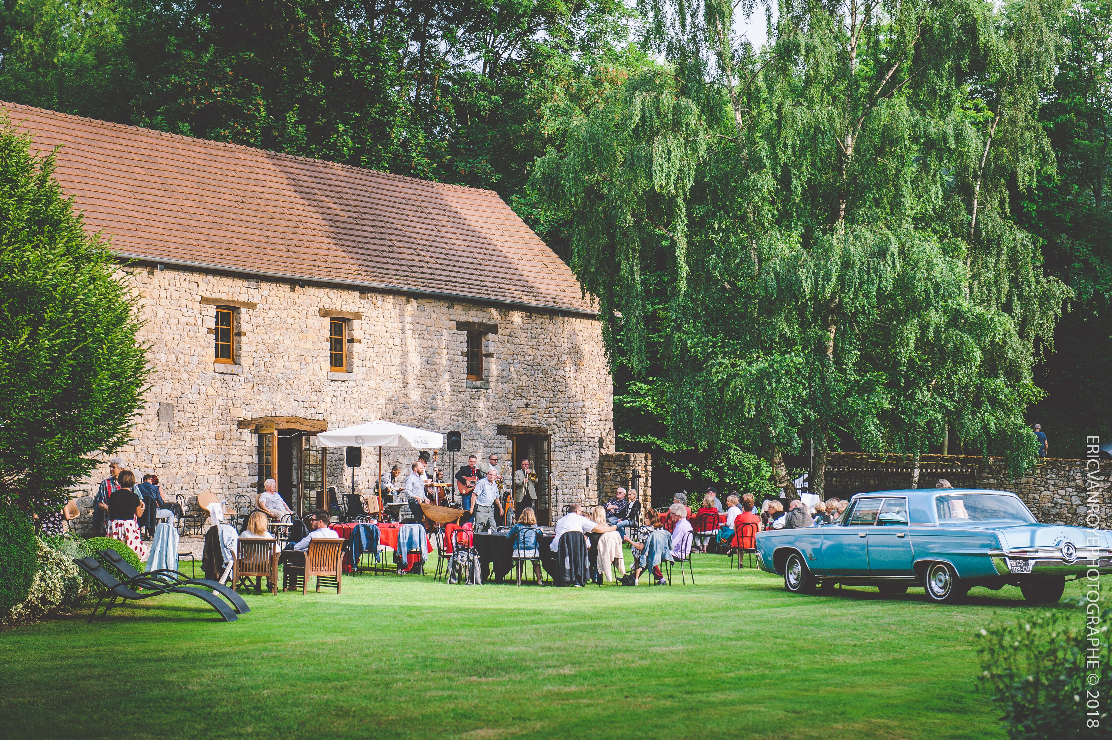 A stone building with a tiled roof, surrounded by lush greenery and a vintage car parked in front.
