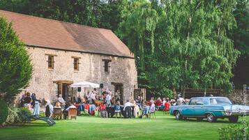 A stone building with a tiled roof, surrounded by lush greenery and a vintage car parked in front.