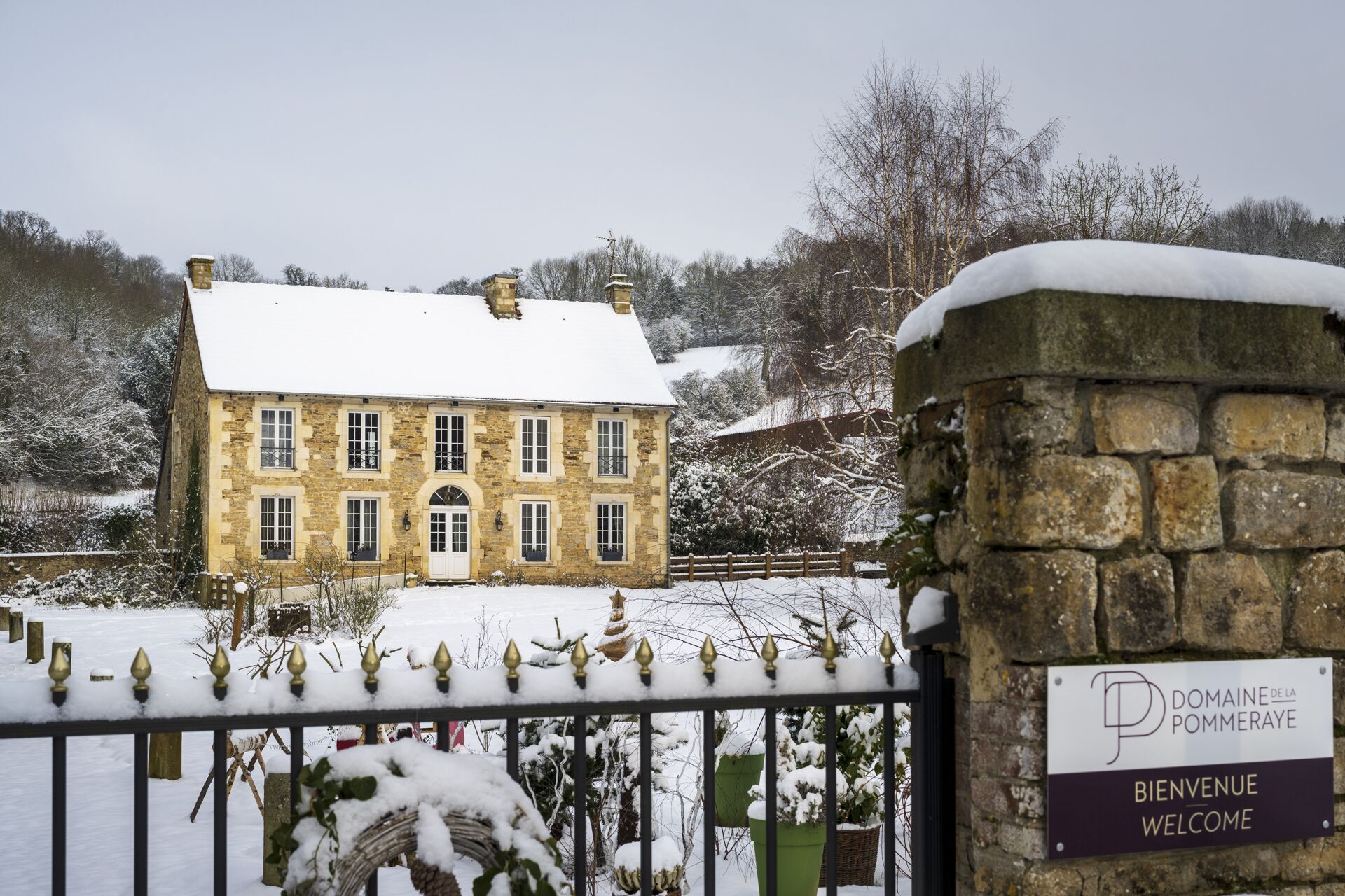 Un bâtiment en pierre avec un toit enneigé, une enseigne indiquant «Bienvenue», et un mur de pierre percé d’une porte.