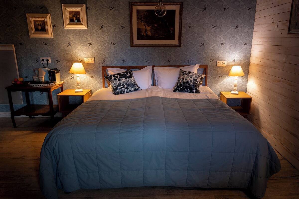 A neatly made bed with a blue quilt, two bedside tables with lamps, and framed pictures on the wall.