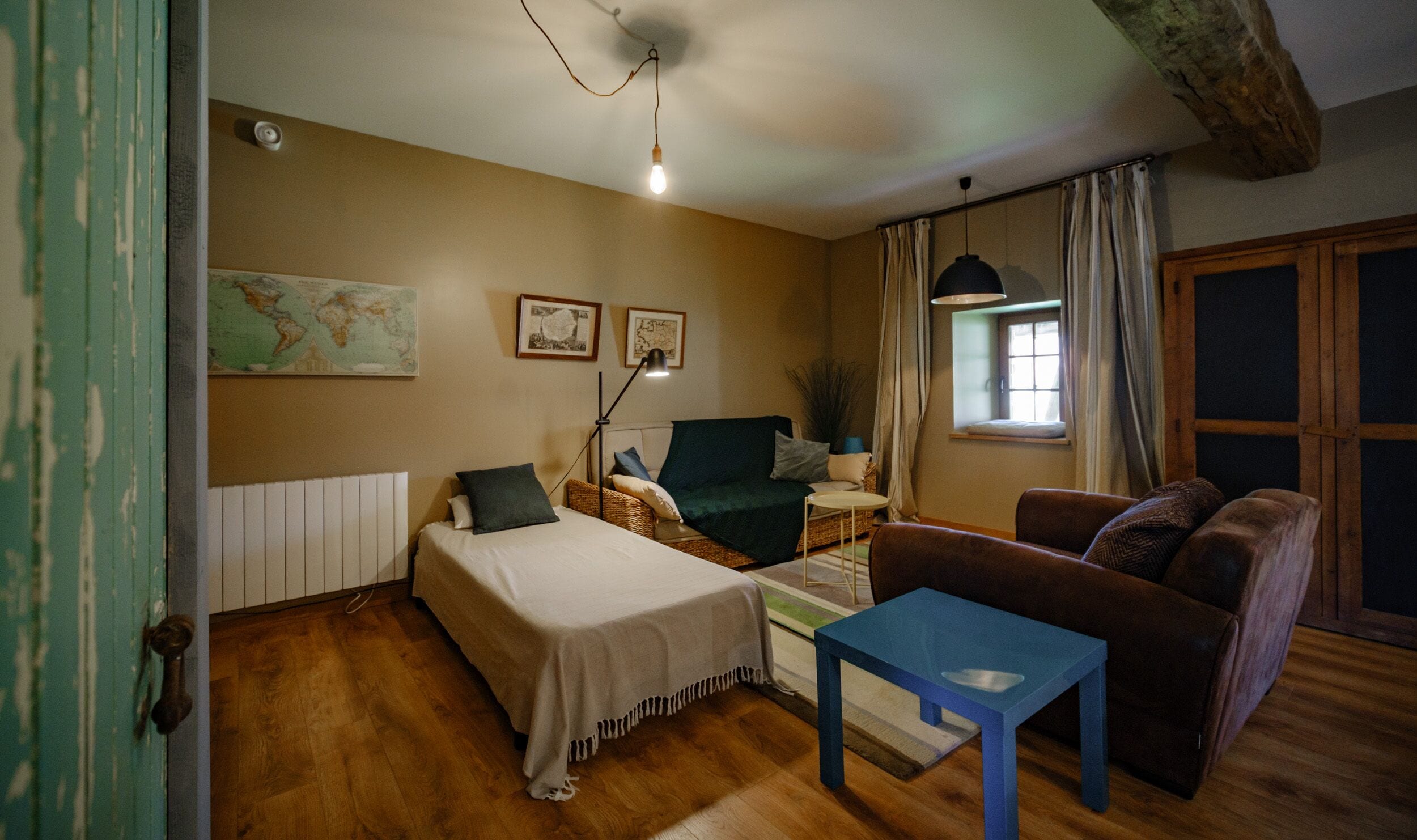 A cozy room with a bed, a blue table, a brown sofa, a map on the wall, and a window with curtains.
