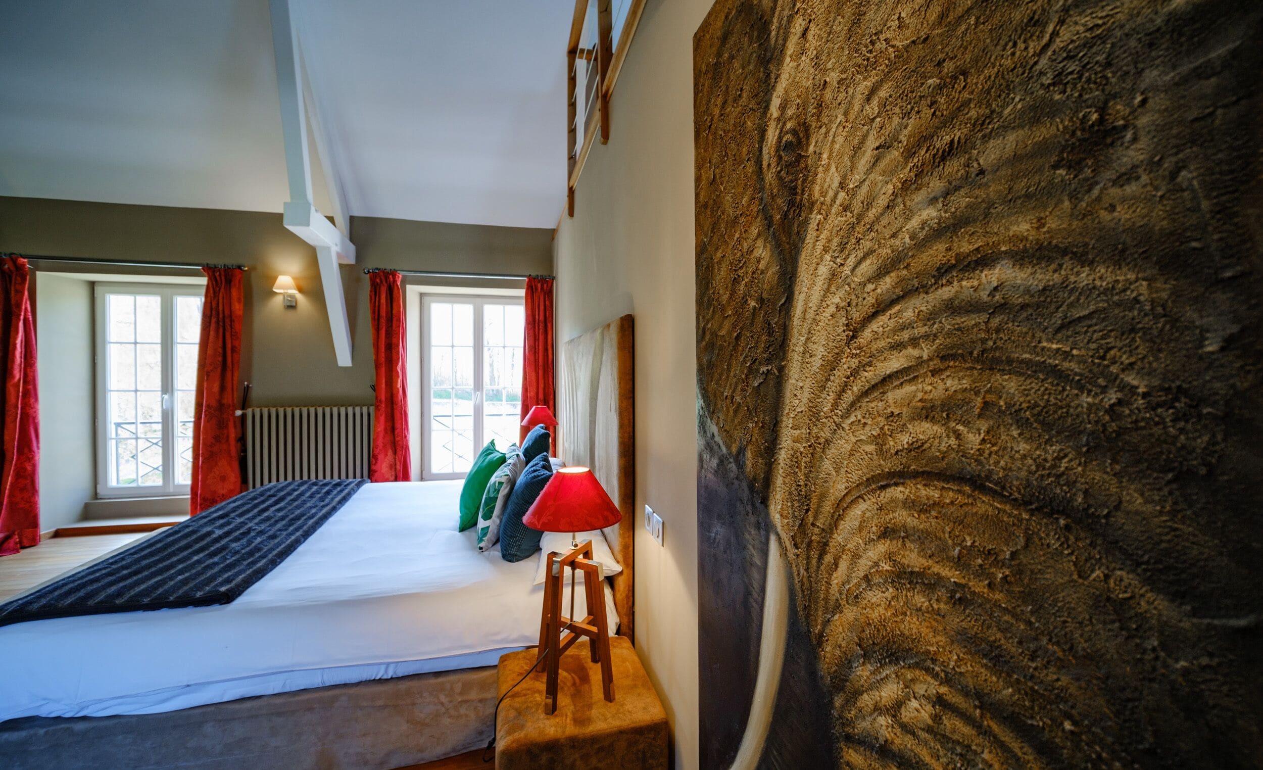 Un dormitorio con una cama, un taburete de madera, una lámpara roja y una pared grande con textura.